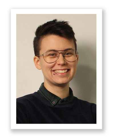 Andy ler stort och tittar in i kamerna. Hen har kort hår, glasögon och en piercing i läppen.